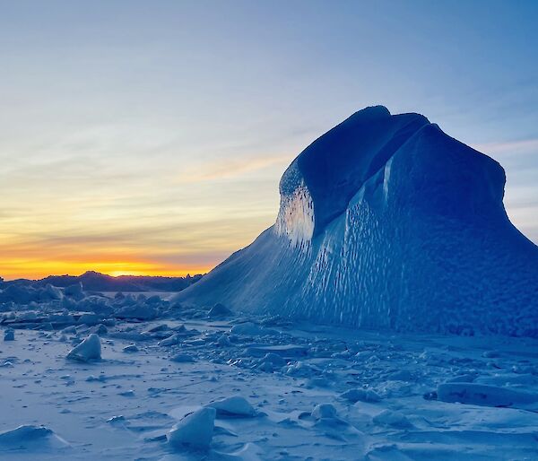 A tall blue iceberg pokes out of the frozen sea-ice. The distant horizon is shades of yellow and orange.
