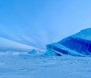 A person in the distance walks across frozen sea-ice towards a tilted dark blue and light blue iceberg..