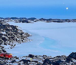 A full moon overhead distant snow-covered hills with a large snow and ice-covered bay in the mid-ground and a red hagg parked on snowy hills in the foreground.