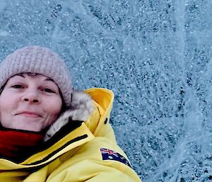 A smiling woman wearing a beanie and yellow jacket takes a selfie with the frozen lake ice behind her.