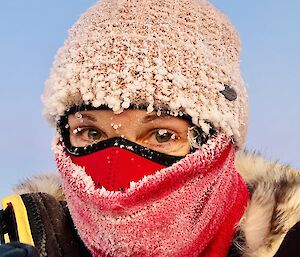 A woman wearing a pink beanie and red buff over her mouth has ice-crystals especially around her eyelashes.