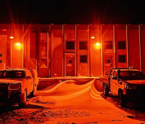 An industrial wokshop with two utes parked out the front on snowy ground is bathed in an orange light from the workshop lights.