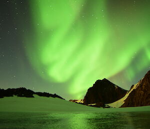 A bright green Aurora lights the sky over a frozen lake with rocky mountain peak in the distance