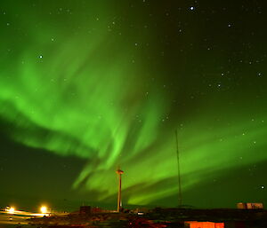 A bright green Aurora lights the sky over a wind turbine with a radio antenna and building on a rocky landscape