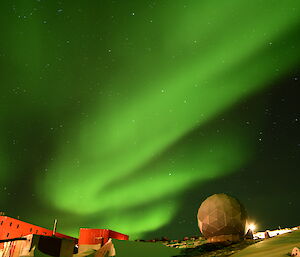 A bright green wavy Aurora is in the sky over bright red buildings on a rocky landscape with a satellite dome in the lower right of frame