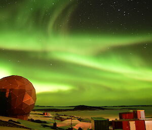 A broad bright green Aurora is in the sky over multi coloured buildings with a satellite dish dome in the lower left of frame