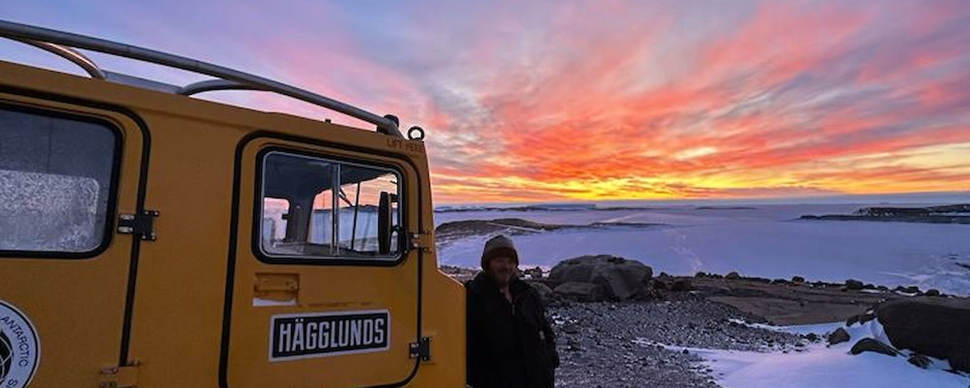 A man is standing next to a yellow Hägglunds with a ice covered harbour and coloured sunset in the distance