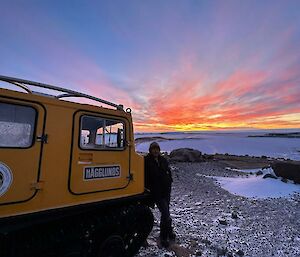 A man is standing next to a yellow Hägglunds with a ice covered harbour and coloured sunset in the distance