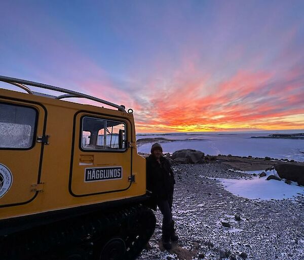 A man is standing next to a yellow Hägglunds with a ice covered harbour and coloured sunset in the distance