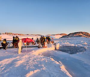 A 3m ractangular hole has been cut in the sea ice and a crowd stands on one side. A red beach umbrella is visible as the sun shines in on a low angle.