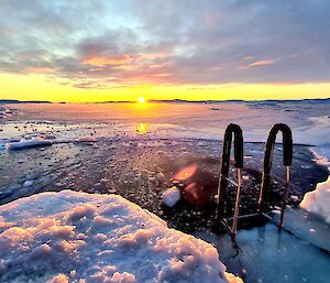 The sun sets, or possibly rises, across a vista of sea ice with a hole carved in the ice in the foreground.
