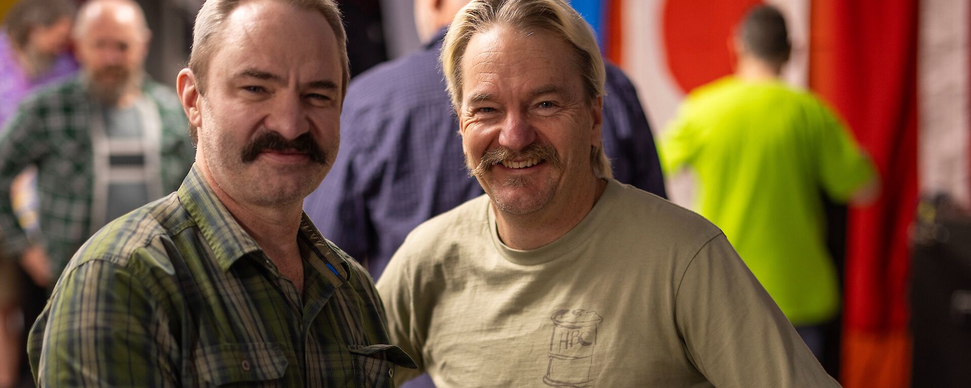 Two moustachioed men smile while holding a small shoebox sized timber box with electronics and lights visible on top.