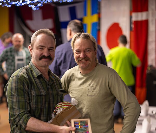 Two moustachioed men smile while holding a small shoebox sized timber box with electronics and lights visible on top.