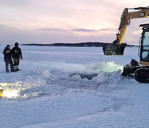 Four men are watching a man use an excavator to remove ice from a hole in the sea ice