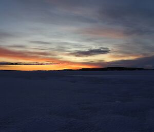 The glow of the sun below the horizon lights the sky over an ice over sea with an island in the distance