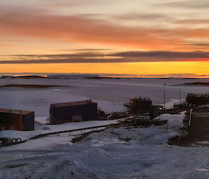 Glow from the sun below the horizon lights the sky in orange in the distance beyond multicoloured buildings on a snow covered shore of a frozen harbour