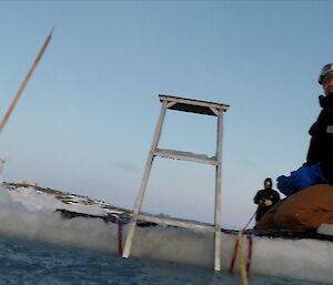 The camera is looking up out of a swim hole in the ice at a man kneeling on the sea ice with a metal ladder in centre of frame