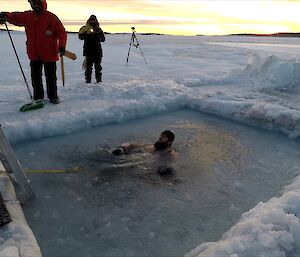 Three men watch on a a man is swimming in icy water inside a hole cut into sea ice