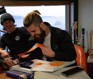 A laughing man opens a gift box containing a fluorescent orange tie. Another laughing man looks on.