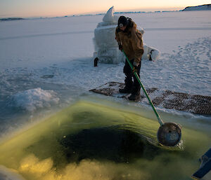 A man uses a colander fixed to the end of a pole to remove sea-ice from the top of a pool cut into the sea-ice.