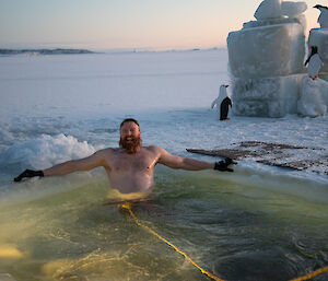 A smiling man reclines in the far corner of a swimming hole cut into the sea-ice.
