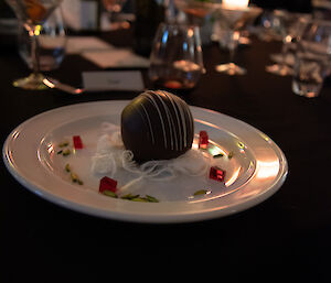 A white plate containing a ball of chocolate mousse coated in hard chocolate, salted caramel, boysenberry pistachio, fairy floss