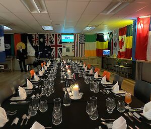 A long dining table with a black tablecloth set for a formal dinner with cutlery, glasses and fancy white napkins. The walls of the room are adorned with international flags.