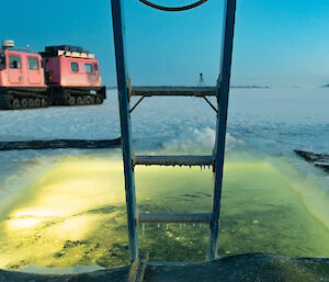 An icicle laden ladder extends into a pool cut into the sea-ice. A large pink vehicle can be seen in the background.