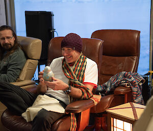 A smiling man wearing a beanie and crocheted multi-coloured scarf admires a hand-made penguin.