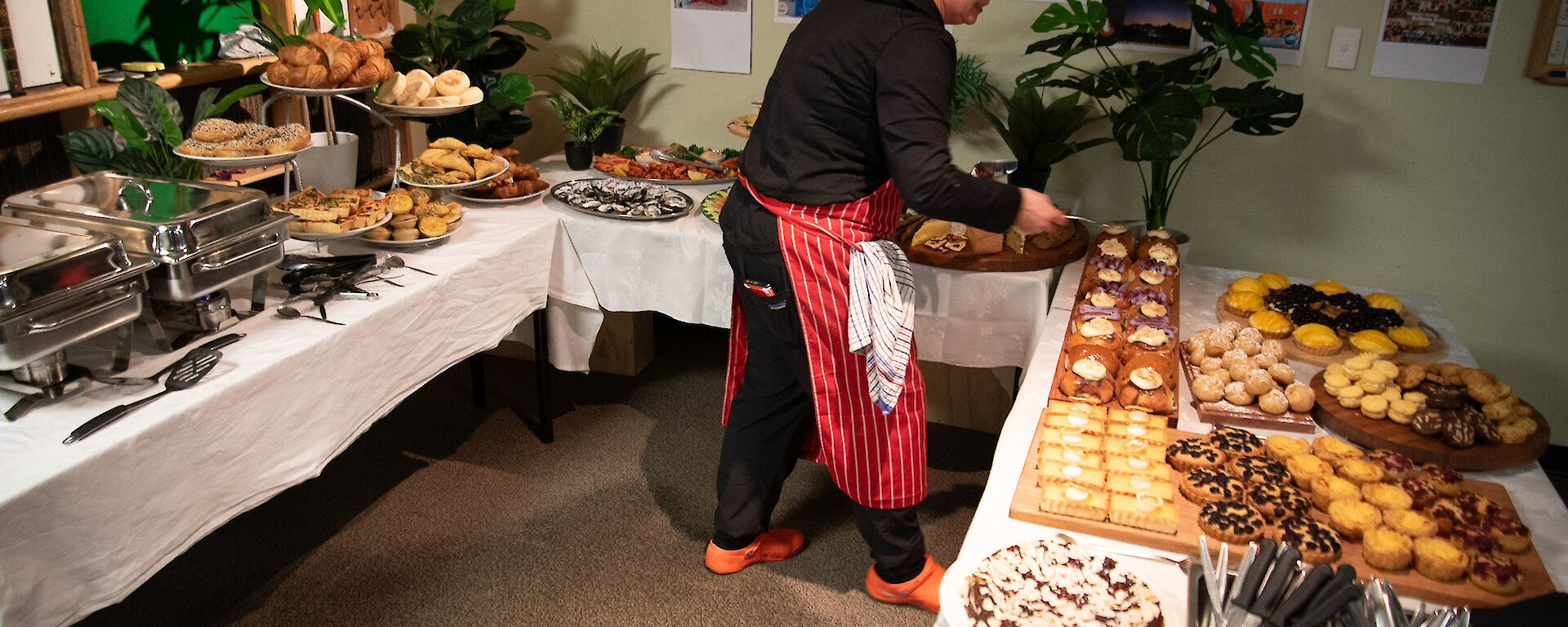 A smiling lady wearing a striped red and white apron lays out food on tables covered in white tablecloths.