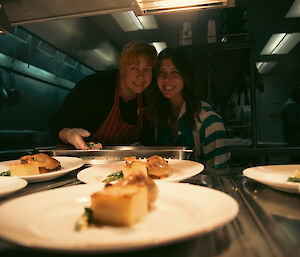 Two smiling women viewed through a bain-marie look at several white plates with food on them.