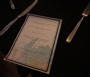 A dinner menu sitting on a black tablecloth between a knife and fork