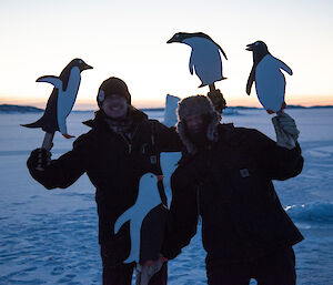 Two laughing men standing on the sea-ice holding hand-made penguin statues.