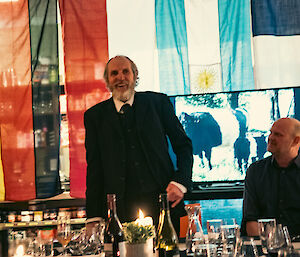 A smiling man standing at the end of a dinner table proposes a toast.