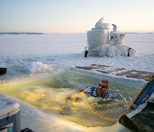 A smiling man wearing a Hawaiin shirt and zinc cream on his nose swims in the swimming hole cut into the sea-ice.