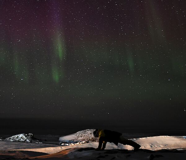 A person undertakes a push up on the snow under a purple and green aurora
