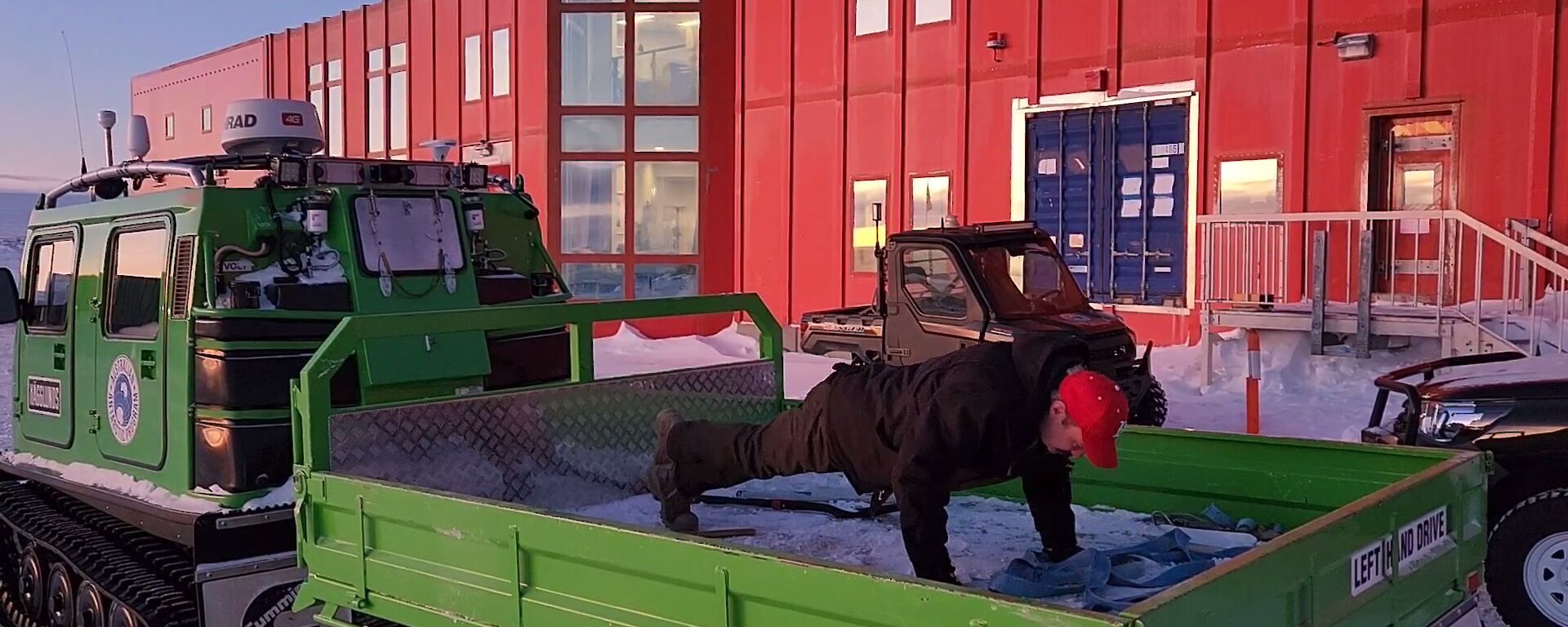 A man performs a push-up in the rear tray of a green tracked vehicle. In the background is a large red two storey building.
