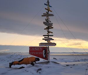 A man performs a push-up on snow covered ground, underneath a sign post. The post is covered from top to bottom in place names, with distances to the places. There is long grey cloud filling the sky across the frame.
