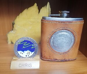 A hand-painted, pewter medallion is in a wooden holder with the name 'Chris' on it. Next to it is a hip flask clad in leather with an emblem embossed