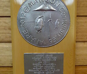 A large plaque with a hand cast pewter motif featuring two penguins, a wind turbine, and a mountain peak, along with a list of names hangs on a wall.