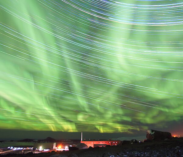 A time lapse photo showing star-trails and a bright green Aurora in the night sky with station buildings on a rocky landscape below.