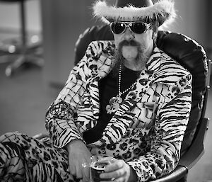 A black and white photo of a man sitting in a chair wearing a gaudy suit jacket and trousers, 'dollar' sign necklace, dark glasses and fur lined cowboy hat.