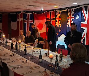 Ten men are sitting, or preparing to sit, at a large table that has been set for a formal dinner. Flags hand in the background and a lighted ice sculpture is in the background.