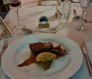 A formal meal with a main course is on a table. A menu is to the top left of frame and a medallion on a personalised stand is in the top centre of frame.