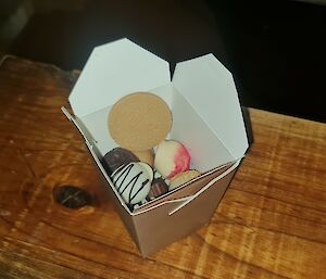 A take-away box filled with different coloured and flavoured chocolate truffles sits on a wooden shelf.