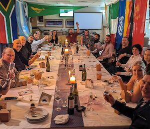 A group of men and women are around a large table having a formal meal. There are flags from many nations hung all around the table.