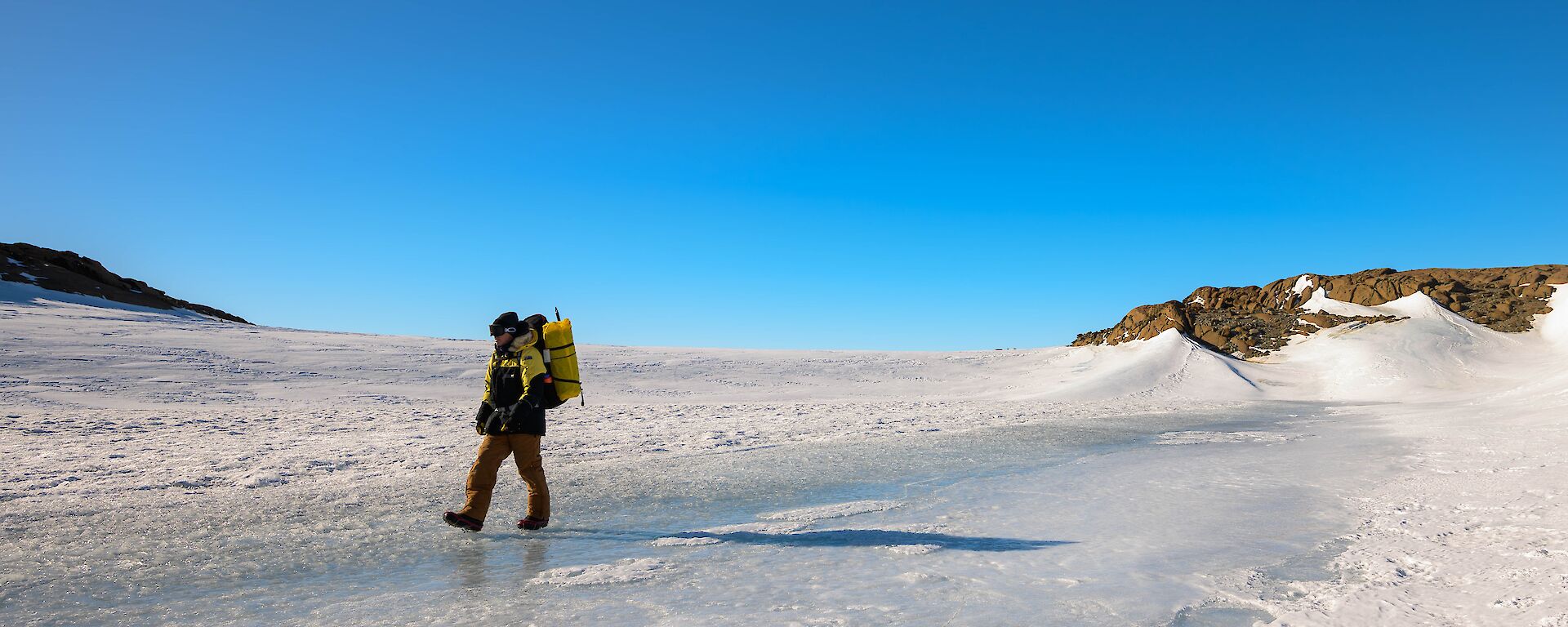 A man in yellow survival clothing, carrying a backpack, is walking across ice covered ground. The sky behind is clear and blue.