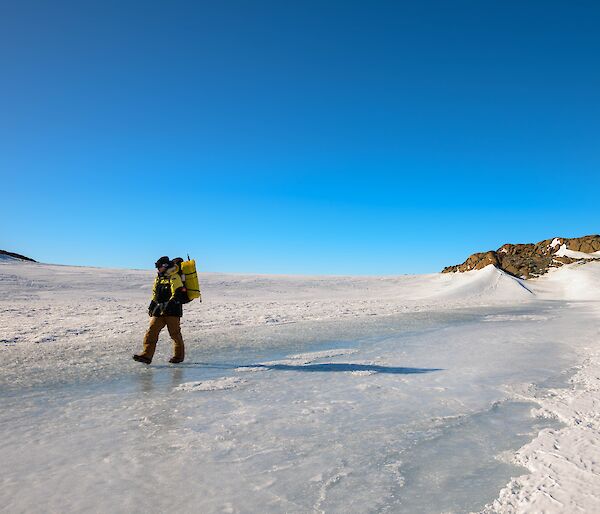 A man in yellow survival clothing, carrying a backpack, is walking across ice covered ground. The sky behind is clear and blue.
