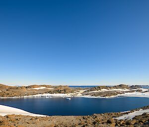 Another landscape photo of a small tranquil bay, surrounded by low lying rocky hills with a light covering of snow. The sky is clear and blue.