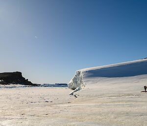 A man to the right of the photo is beginning to walk up a snow covered hill. In the centre of the photo, the hill has become an ice cliff face, with the thin sea ice at its' base. It is a sunny day with a clear sky.